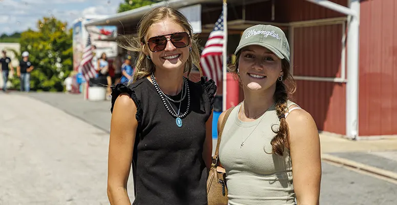 Two women posing together at an outdoor fair on a sunny day, smiling; one wears a black sleeveless top and sunglasses, the other a gray tank and cap, with US flags in the background.