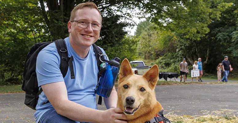 Man in a blue shirt with a backpack and water bottle sits beside a tan dog wearing a vest at a park event.