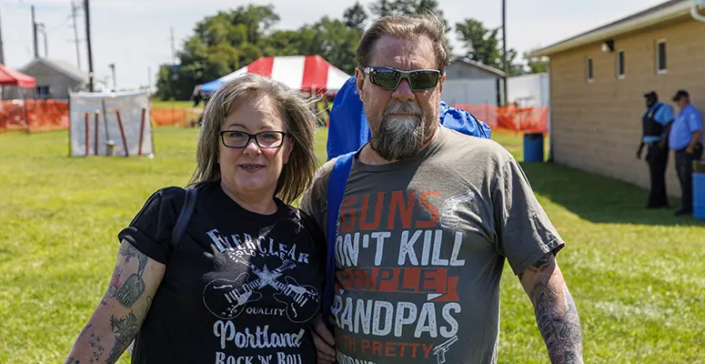 Smiling man and woman wearing sunglasses stand together at an outdoor event on a sunny day, with tents and orange fencing in the background.