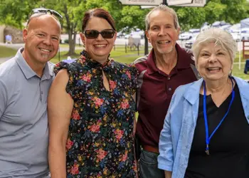 Four adults smiling and posing outdoors at a park event, standing side by side (two men on sides, two women in the middle).