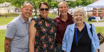 Four adults smiling and posing outdoors at a park event, standing side by side (two men on sides, two women in the middle).