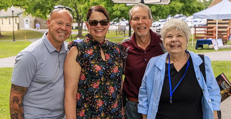Four adults smiling and posing outdoors at a park event, standing side by side (two men on sides, two women in the middle).