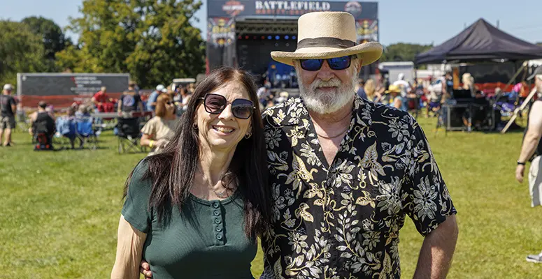 Smiling man with a straw hat and beard wearing a black floral shirt stands next to a smiling woman in a green top at an outdoor festival; stage and tents in the background.