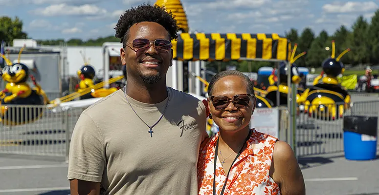 Two smiling people wearing sunglasses stand together at a fairground with a yellow bee-themed ride in the background.