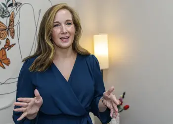Woman in a blue wrap dress speaks with open hands in a room with butterfly mural and a lamp behind her.