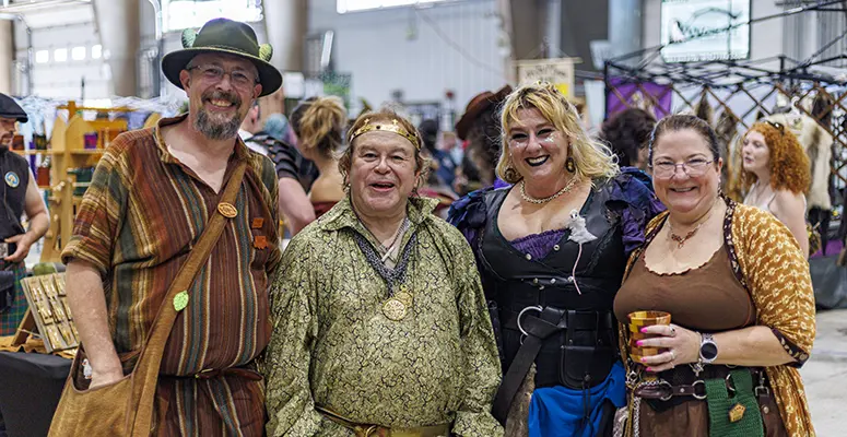 Four adults in medieval costumes posing together at an indoor festival, smiling at the camera.
