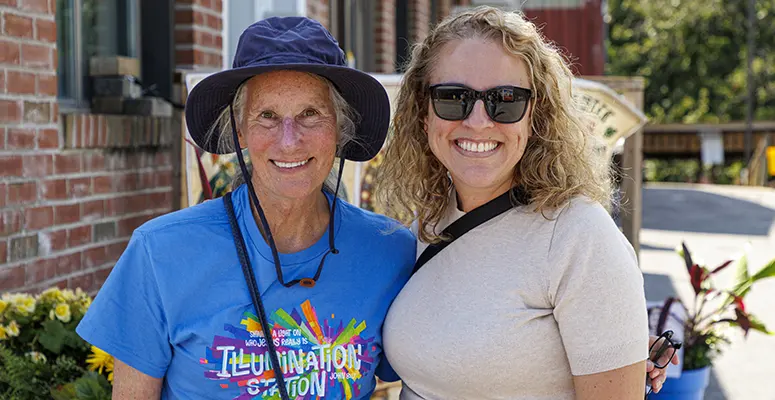 Two smiling women pose outdoors next to a brick building; one wears a blue hat and a colorful graphic T‑shirt, the other a light beige top and dark sunglasses.