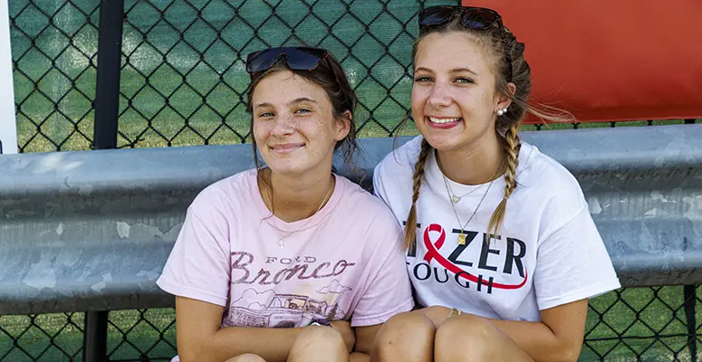 Two girls sit side by side on a metal barrier at an outdoor sports field, smiling at the camera.