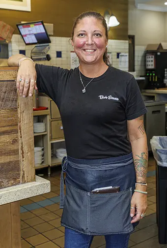 Smiling female Bob Evans employee in a black shirt and blue apron stands behind a restaurant counter.