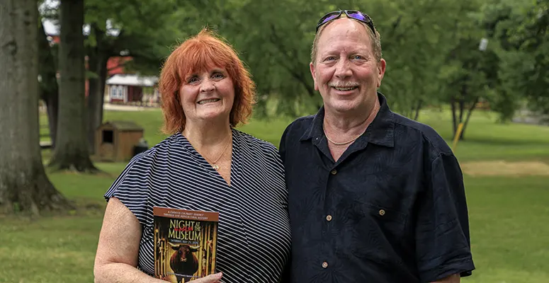 Smiling couple posing in a park; the woman holds a flyer for a museum event that reads 'Night at the Museum'.