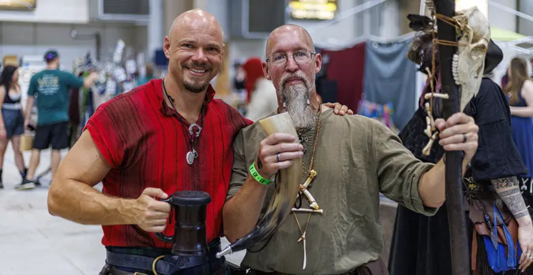 Two men in medieval cosplay posing together at a convention, one in a red tunic with a mug and the other with a long beard holding a horn and a staff.