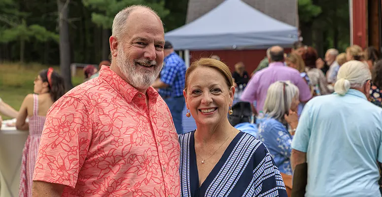 Smiling couple at an outdoor gathering, man in a coral floral shirt and woman in a blue striped blouse, with attendees and a canopy in the background.