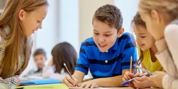 Group of four kids sit at a table, writing and drawing together in a bright classroom, smiling and helping each other on a project.