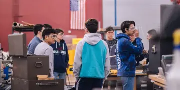 Group of students in a machine shop standing around workbenches and equipment, discussing a project, with an American flag in the background.