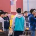 Group of students in a machine shop standing around workbenches and equipment, discussing a project, with an American flag in the background.