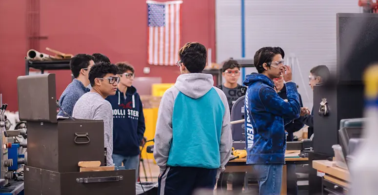 ccps-career-tech-image - Carroll News | Carroll Publications | Carroll County, MD Group of students in a machine shop standing around workbenches and equipment, discussing a project, with an American flag in the background.