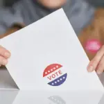 Hands hold a white ballot card with a red, white, and blue 'VOTE' sticker on a table.