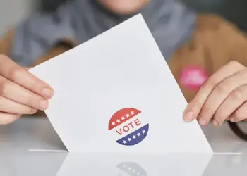 Hands hold a white ballot card with a red, white, and blue 'VOTE' sticker on a table.