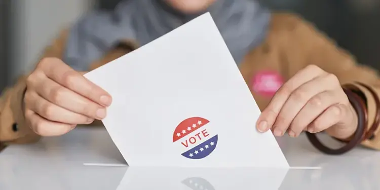 Hands hold a white ballot card with a red, white, and blue 'VOTE' sticker on a table.