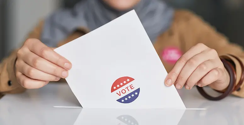 Hands hold a white ballot card with a red, white, and blue 'VOTE' sticker on a table.