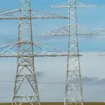 Metal lattice transmission towers carrying high‑voltage lines across a flat field under a blue sky.