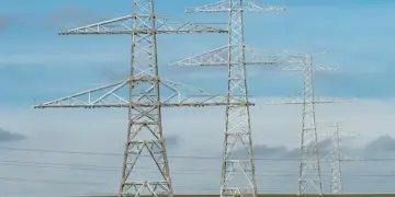 Metal lattice transmission towers carrying high‑voltage lines across a flat field under a blue sky.