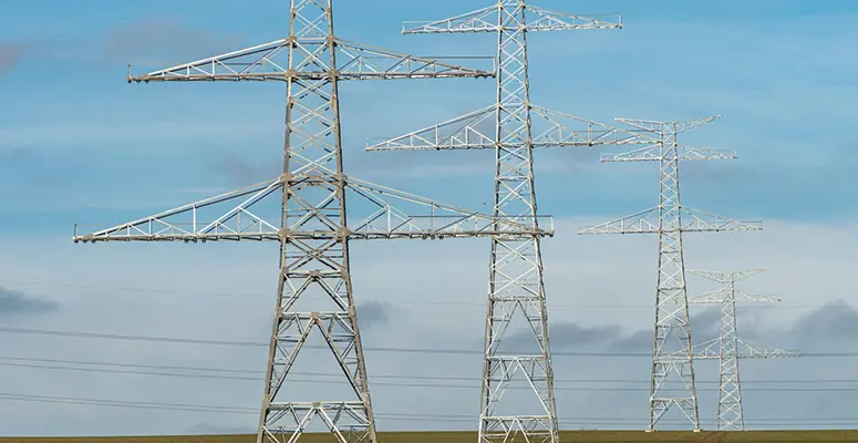 Metal lattice transmission towers carrying high‑voltage lines across a flat field under a blue sky.
