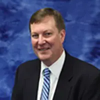 Head-and-shoulders portrait of a smiling man in a dark suit and striped tie against a blue studio backdrop.