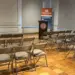 Empty conference room with rows of brown folding chairs facing a podium and banner at the front of the room.