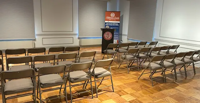 Empty conference room with rows of brown folding chairs facing a podium and banner at the front of the room.