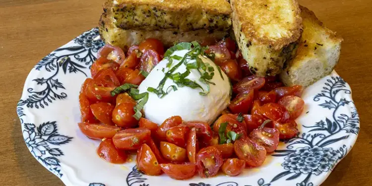 Plate of chopped cherry tomatoes around a burrata ball, topped with basil, with toasted bread slices on a blue-and-white patterned plate.