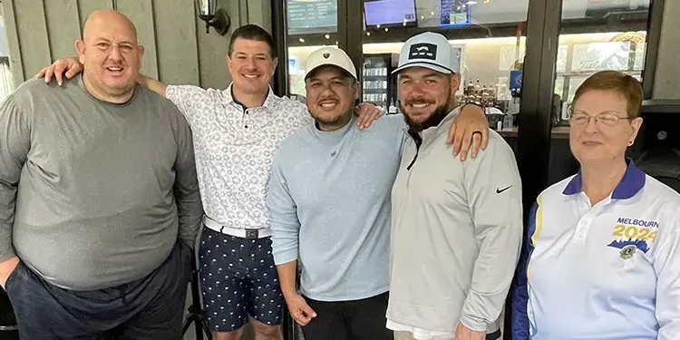 Five adults posing with arms around each other in a cafe, smiling at the camera.