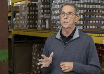 Older man wearing glasses speaks and gesticulates in a warehouse aisle with shelves of boxed goods behind him, suggesting a briefing or tour.