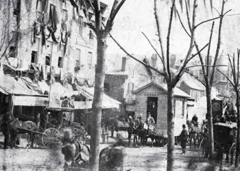 Historic city street with horse-drawn carriages, vendors, and pedestrians along a row of shops on a tree-lined avenue.