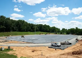 Construction site for a sports field in progress: a large excavated area lined with black tarp, with several trucks and equipment and dirt mounds around it, trees in the background.