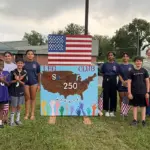 Group of children and adults posing beside a handmade sign reading LEO Club USA 250, with an American flag overhead.