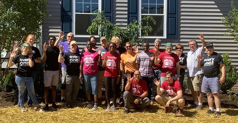 Group of volunteers posing outside a gray building, many waving at the camera. (Informative group photo)